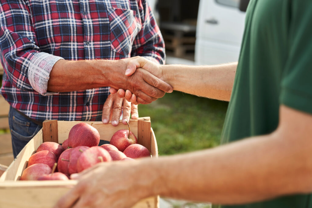 Hands of a grower and supplier shaking hands over a crate of fresh apples, representing local Pacific Northwest fruit sourcing.