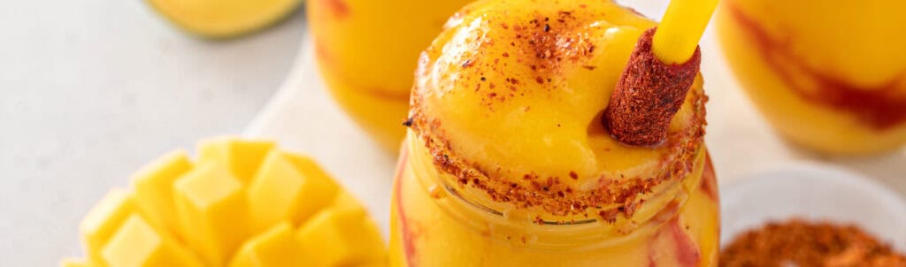 Mango chamoy drink in glass jar with chili powder rim and tamarind straw highlighting the swicy flavor trend. Mangonada mexican mango smoothie with chamoy sauce and chili lime seasoning and tamarind candy straw