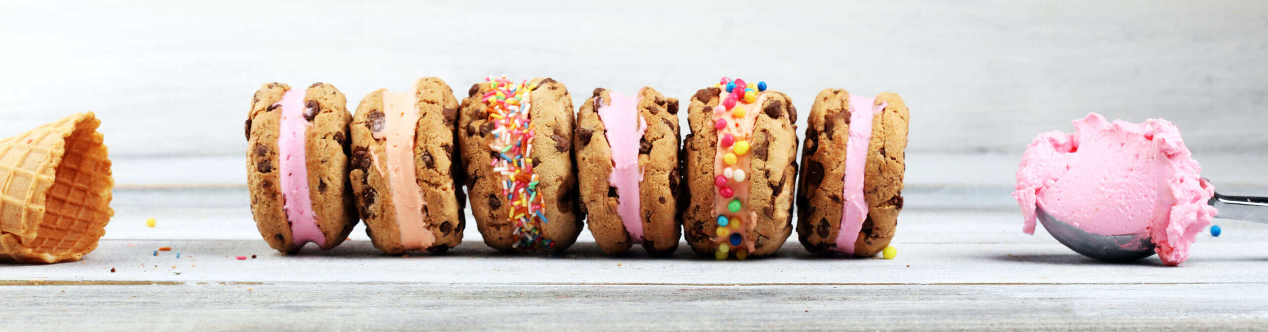Row of chocolate chip cookie ice cream sandwiches with pink ice cream filling and colorful sprinkle variations, demonstrating format innovation and nostalgic treat reimagining with modern toppings