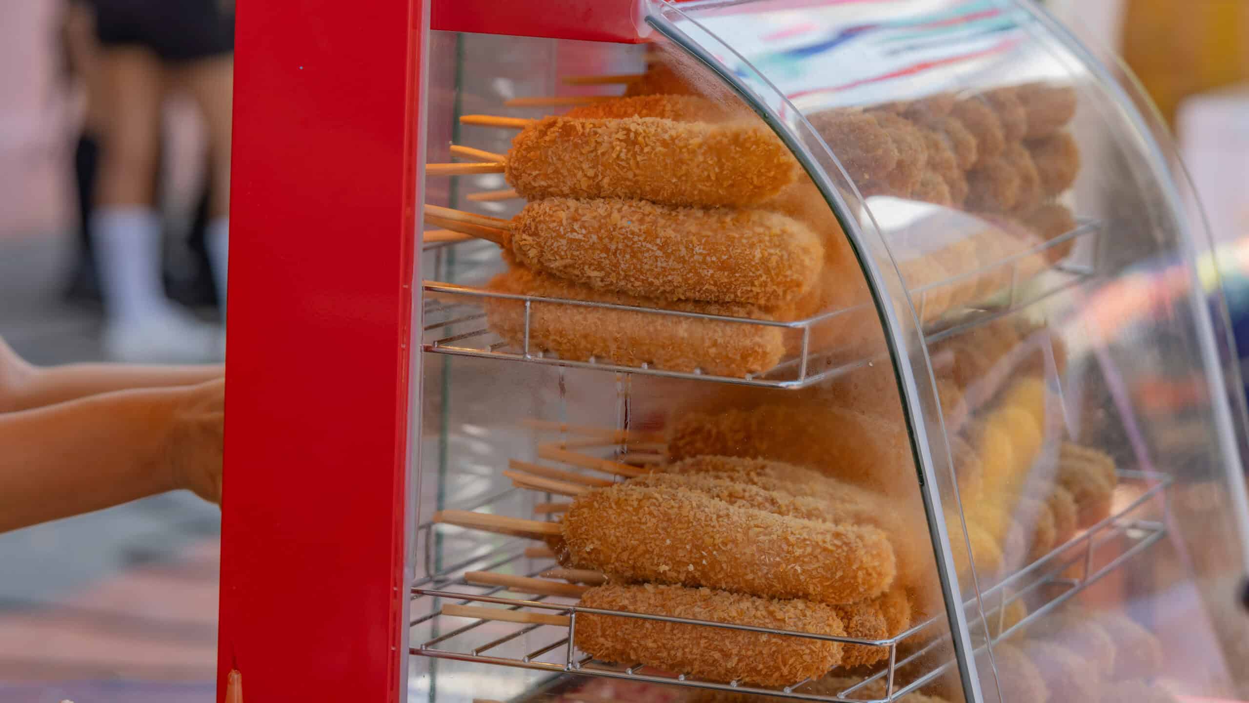 Crispy fried corn dogs on wooden sticks stacked in a heated red street food display case