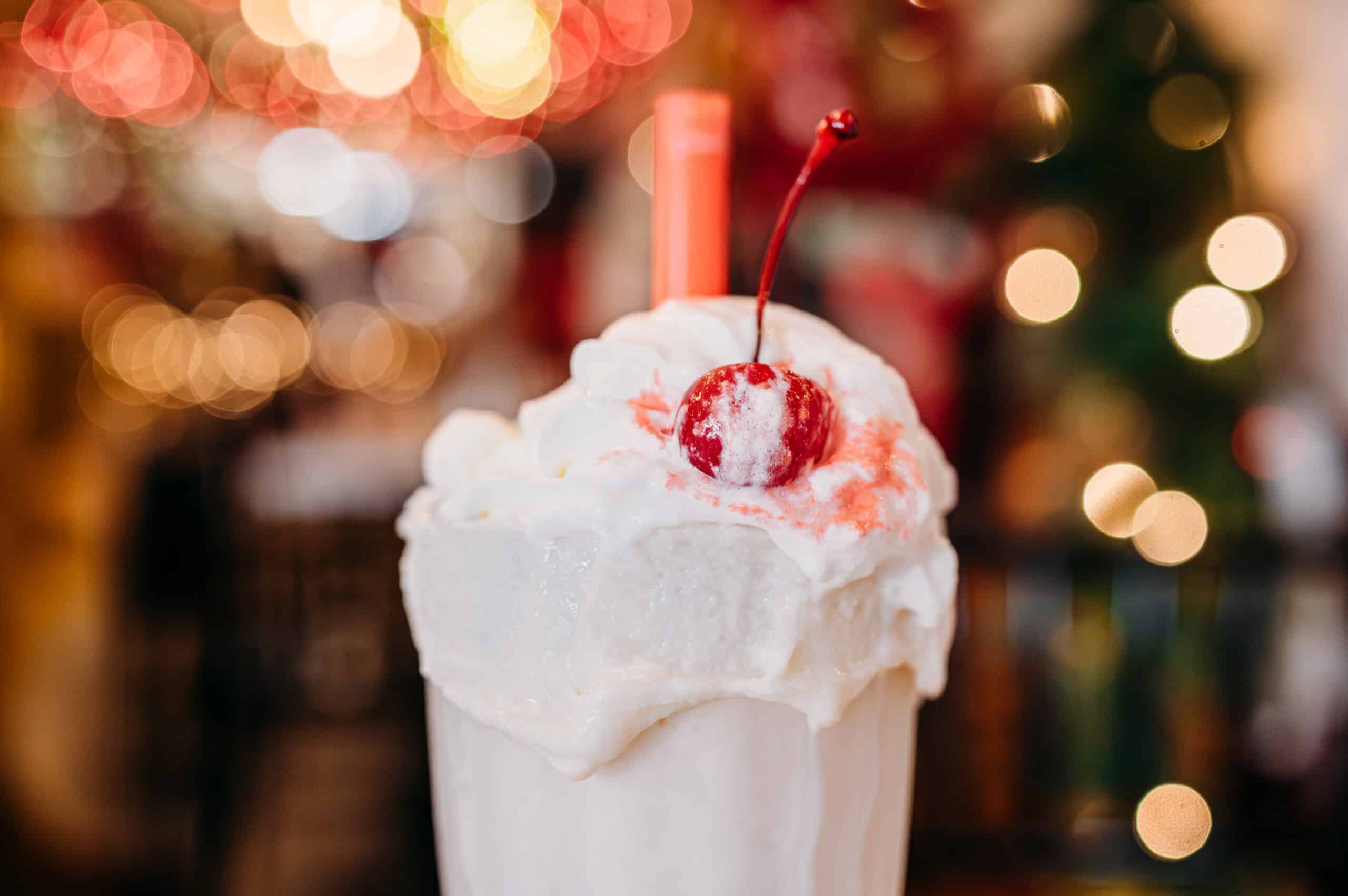 A nostalgic creamy vanilla flavor milkshake overflowing with whipped cream and topped with a maraschino cherry in a warmly lit retro diner