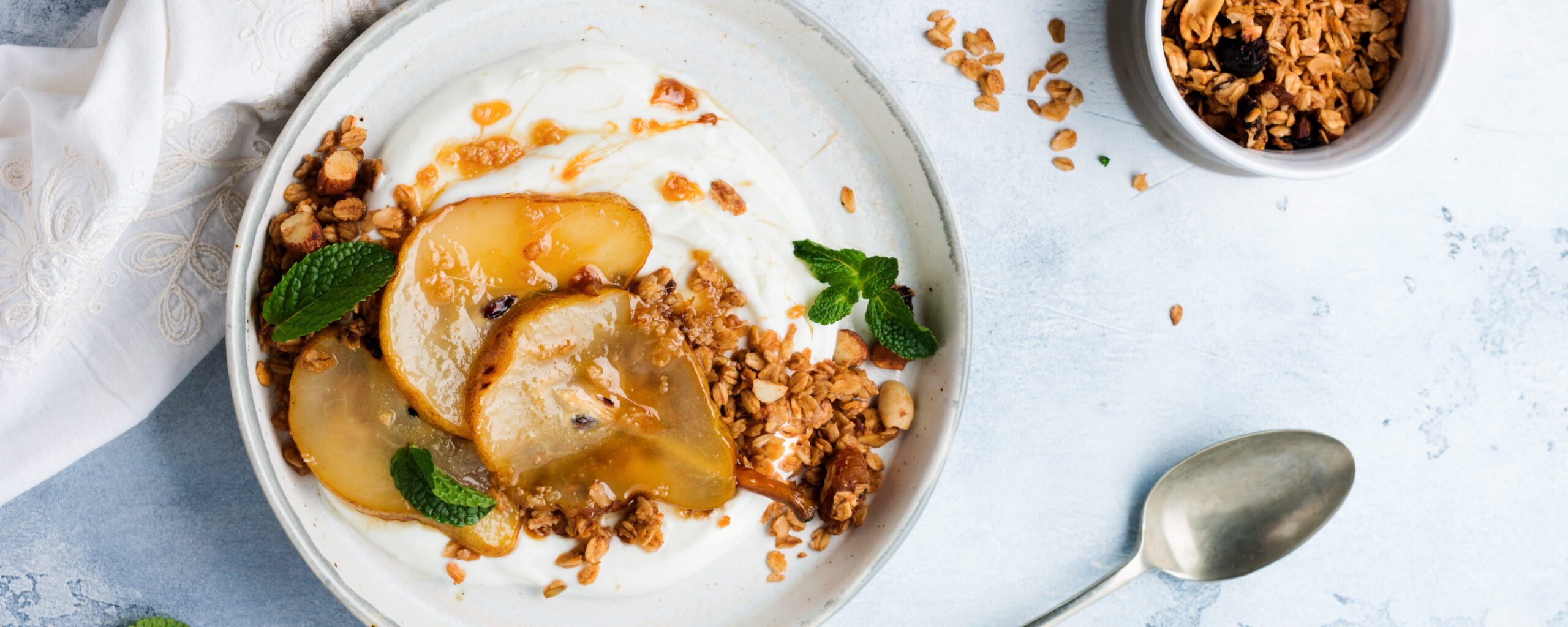 Overhead view of premium yogurt bowl with caramelized pear slices, granola, hazelnuts, and fresh mint on a light blue surface representing the culmination of cultured dairy product development trends
