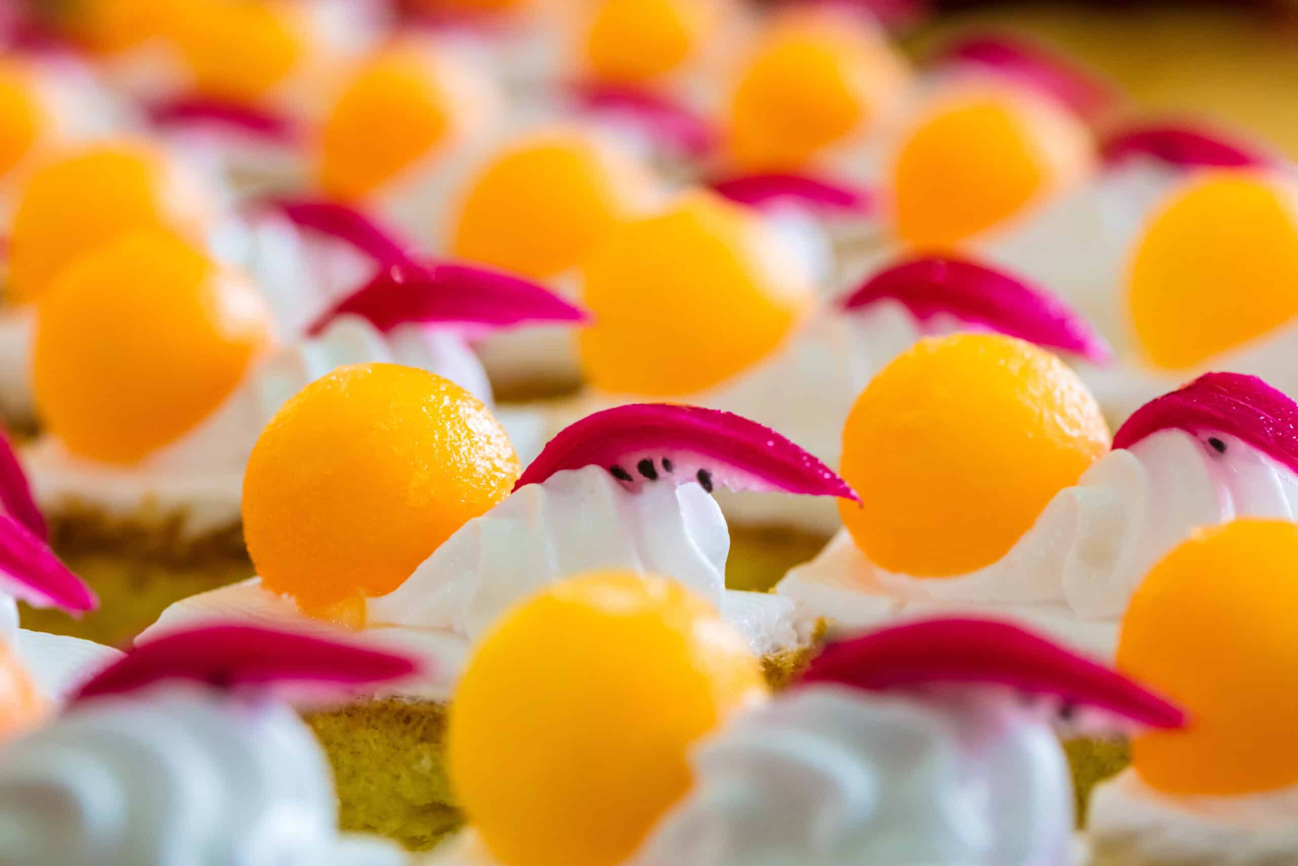 Rows of artisanal petit fours topped with mango spheres and dragon fruit slices showcasing tropical fruit trends in bakery