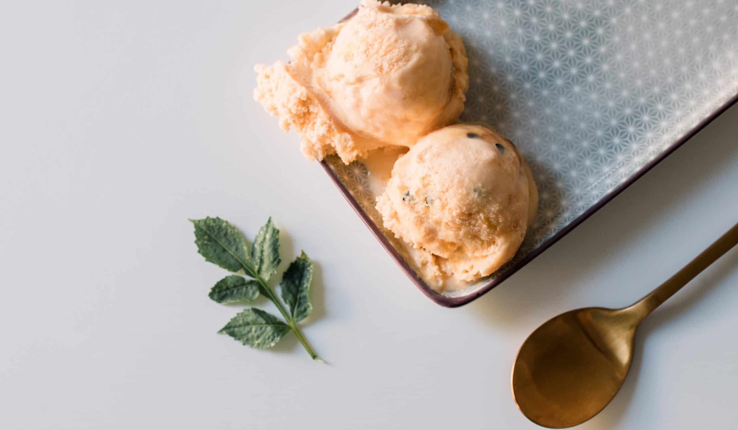 Peach ice cream scoops in a patterned tray with mint and a brass spoon — premium styling reflecting rising ingredient costs in ice cream formulation.