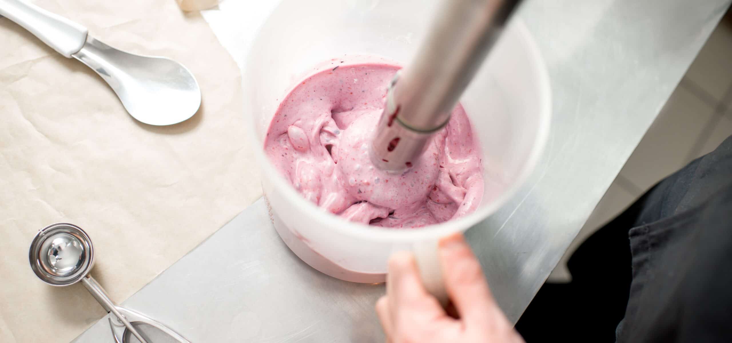 Hands blending a berry ice cream base in a small-batch development kitchen — the processing stage where ingredients must hydrate and disperse evenly.
