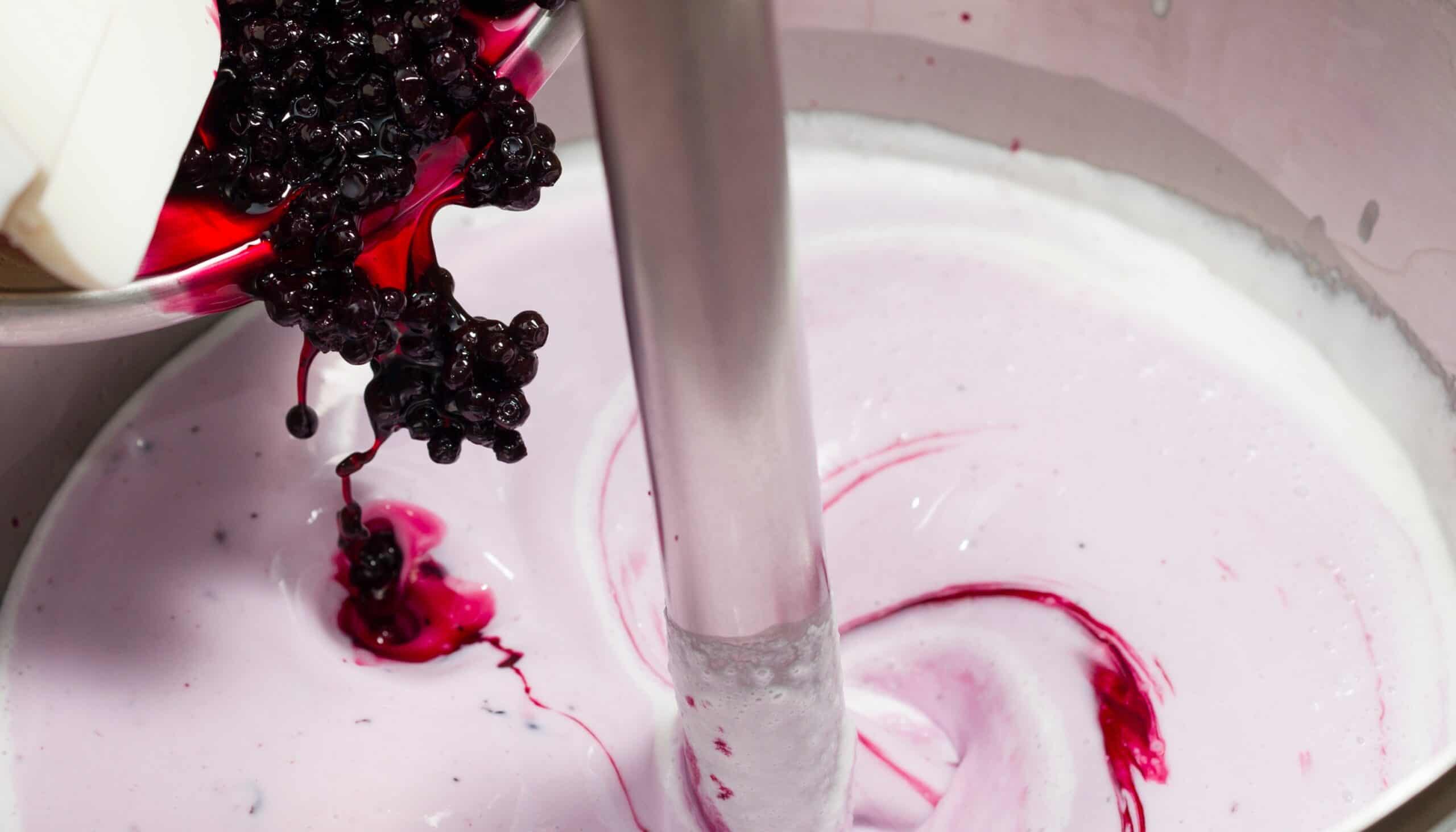 Blackberries pouring from a metal scoop into a pink ice cream base — the processing stage where functional and botanical ingredients are incorporated.