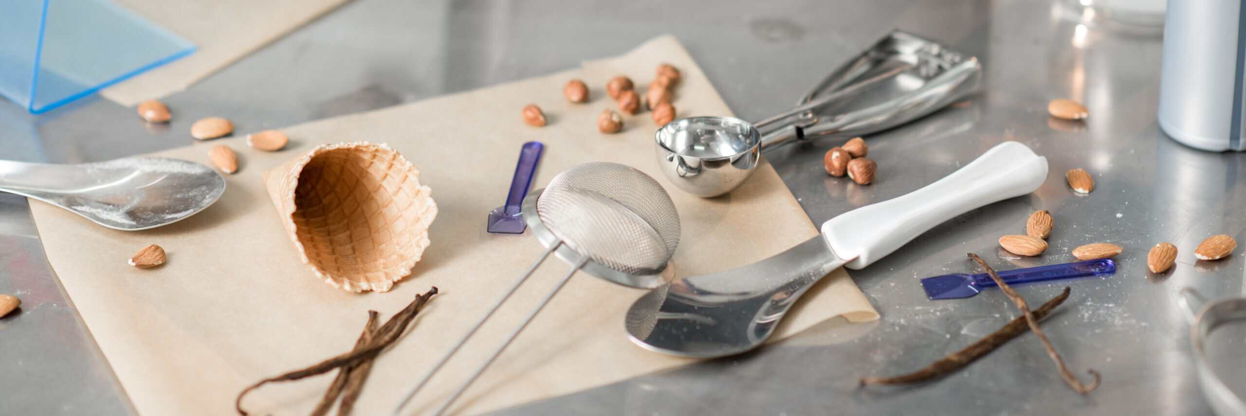 Vanilla beans, almonds, hazelnuts, waffle cone, and stainless tools scattered on parchment — the R&D bench behind ice cream development.