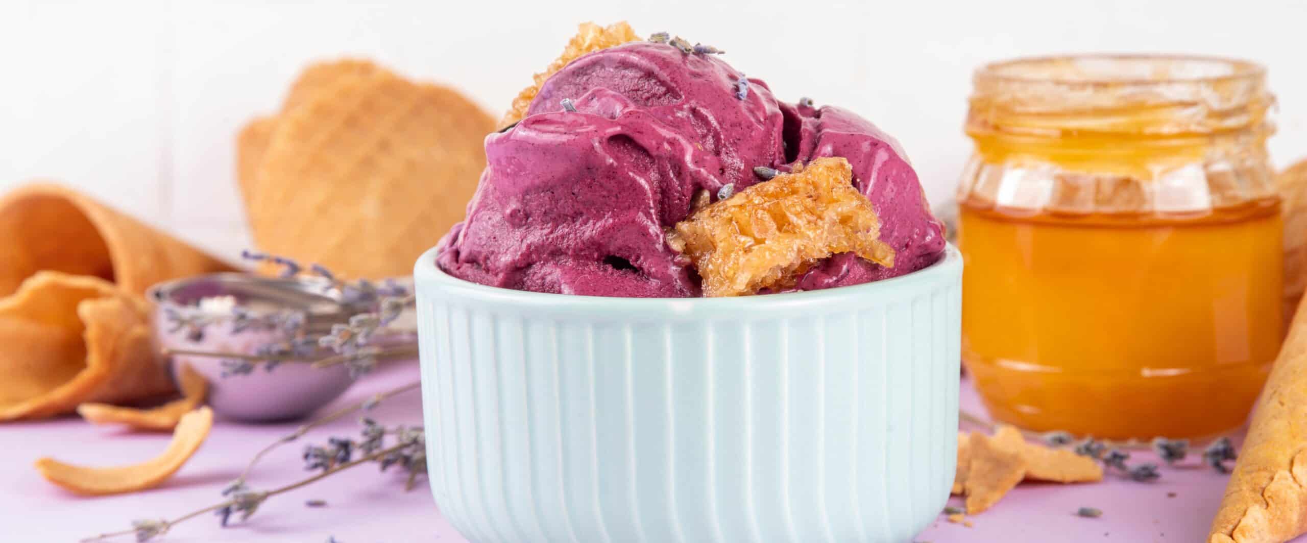 A delicious scoop of berry-flavored ice cream in a white bowl, garnished with cookie pieces, with waffle cones and honey jar in the background, perfect for summer treats.