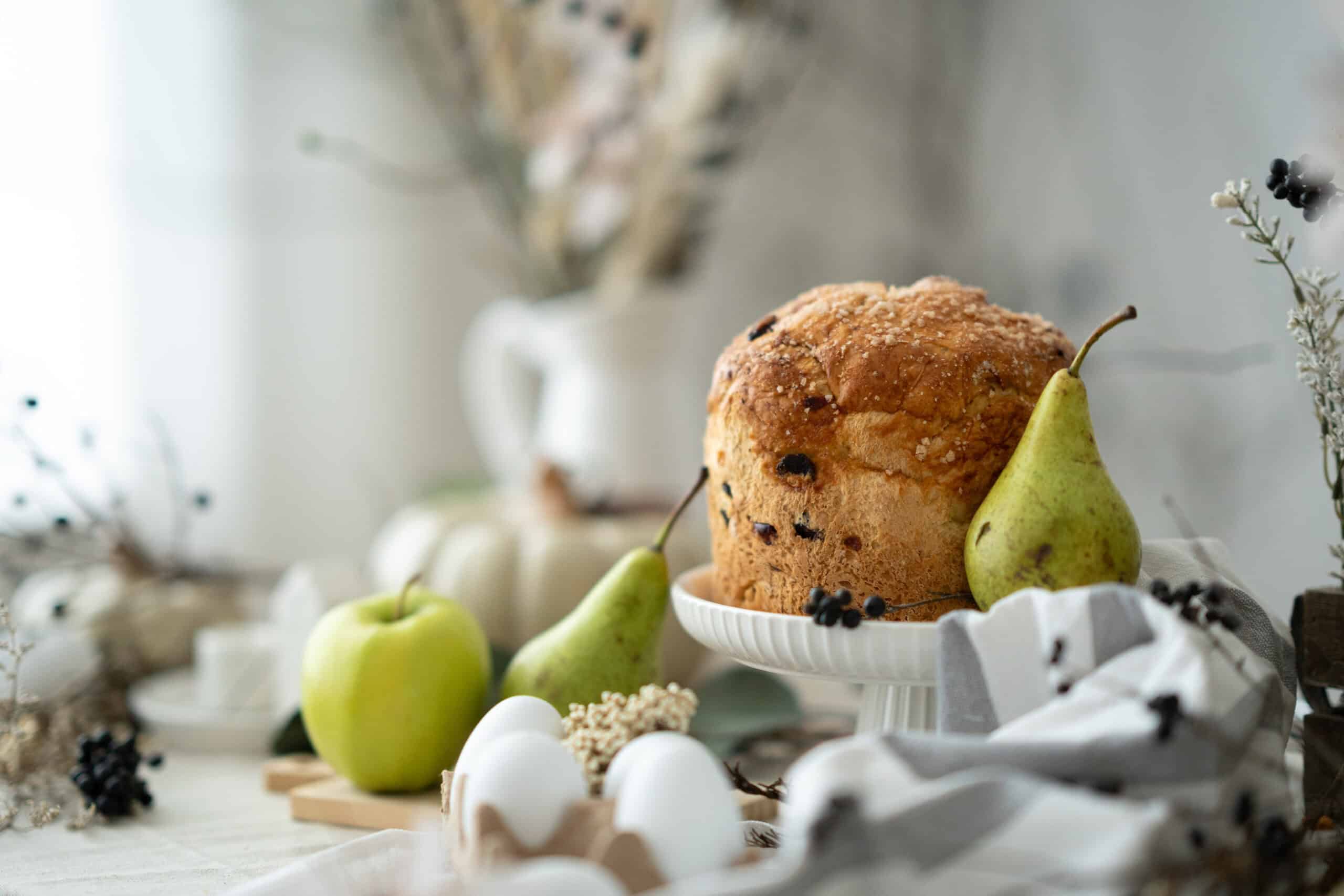 Rustic panettone on a white cake stand surrounded by green pears, apples, white pumpkins, and dried botanicals, evoking autumn botanical flavors in bakery.