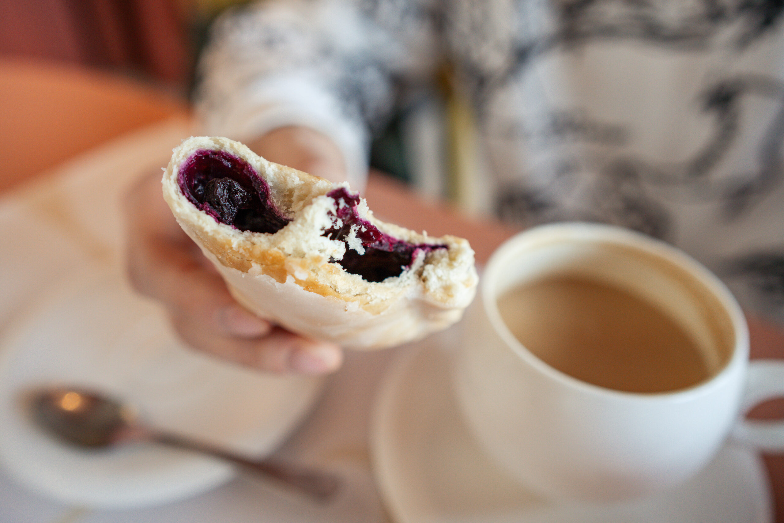 A persons hand holding a halved blueberry hand pie beside a cup of coffee, revealing soft bake-stable fruit filling inside.