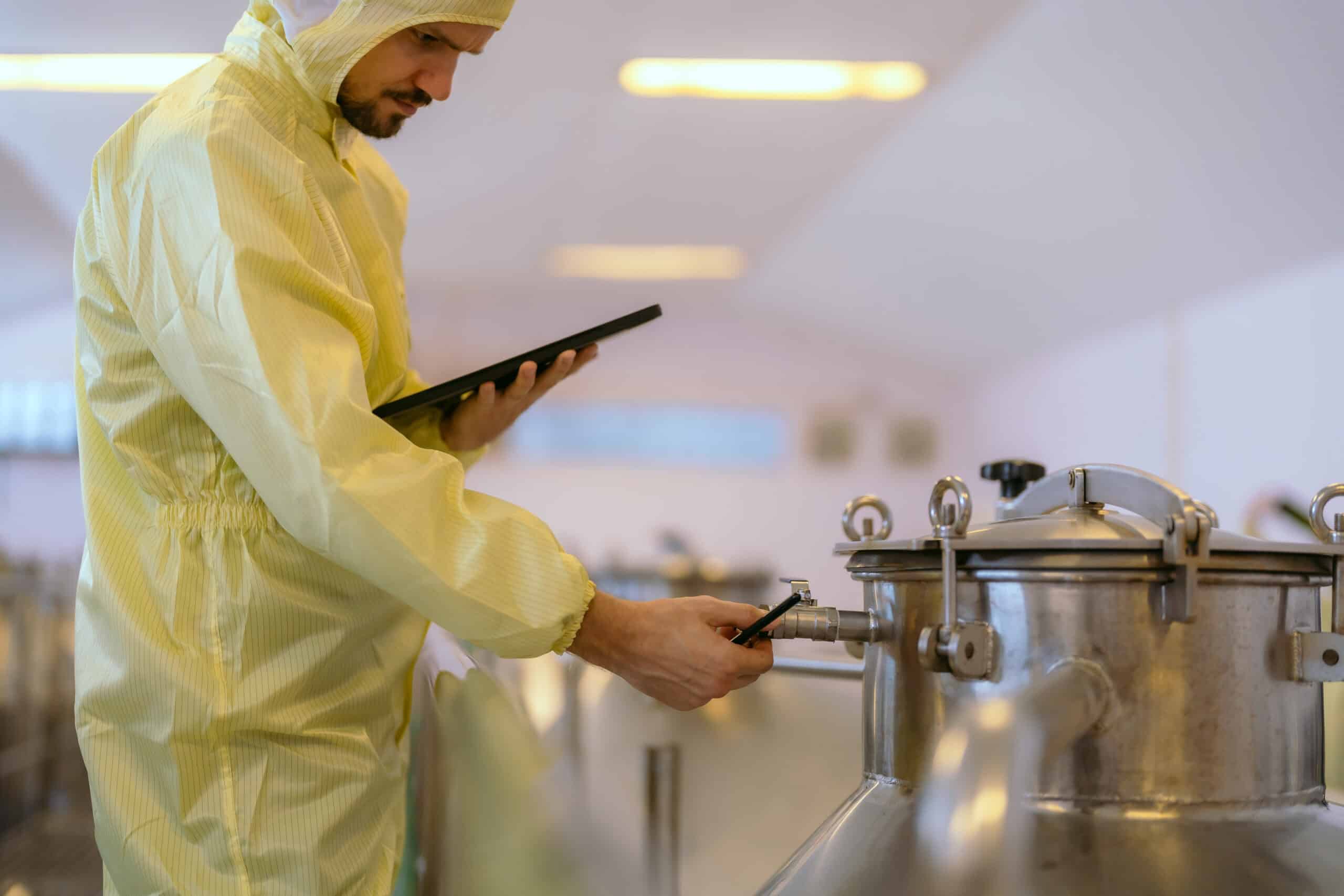 Inspector in a protective gown and blue gloves recording checks on a clipboard beside food production equipment during a ingredient quality GMP audit.