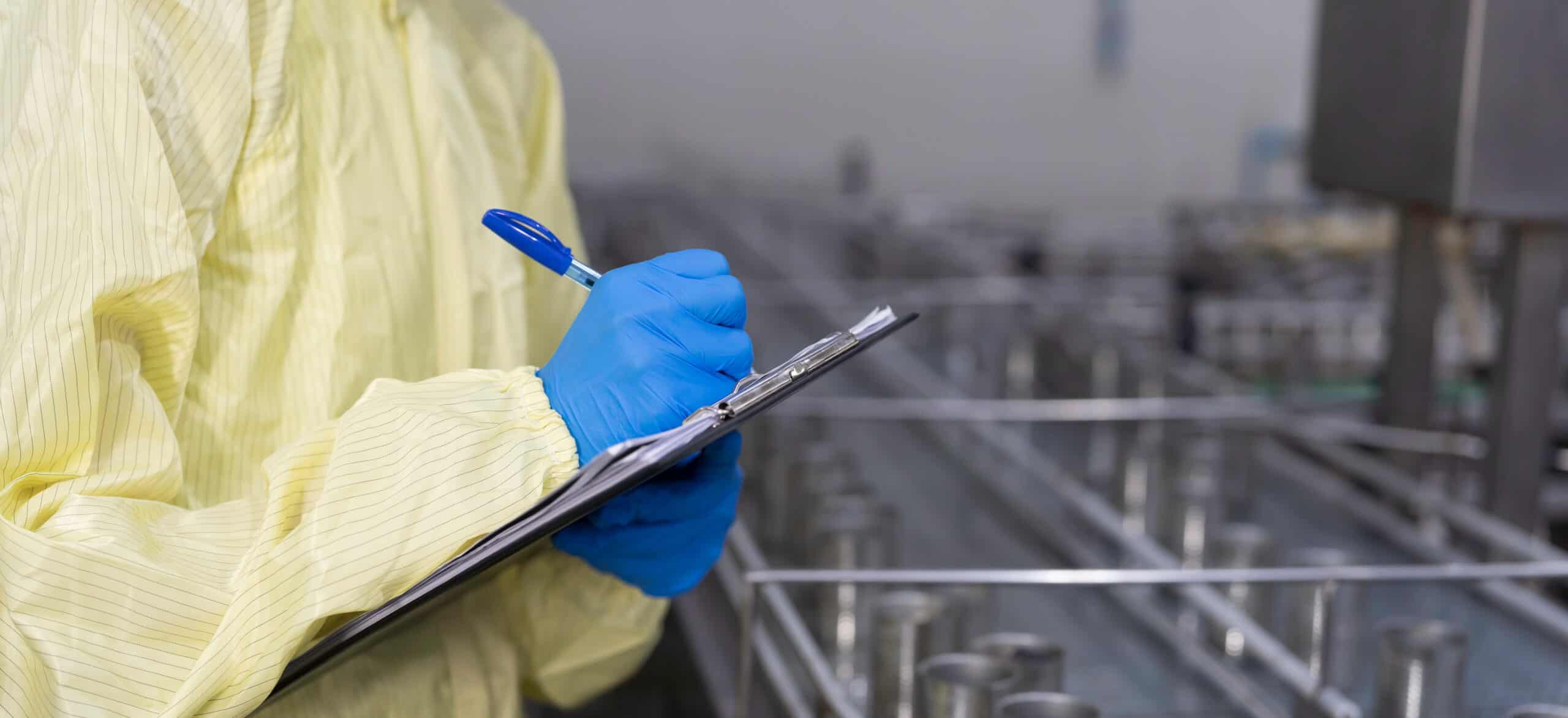 Inspector in a protective gown and blue gloves recording checks on a clipboard beside food production equipment during a GMP quality audit.