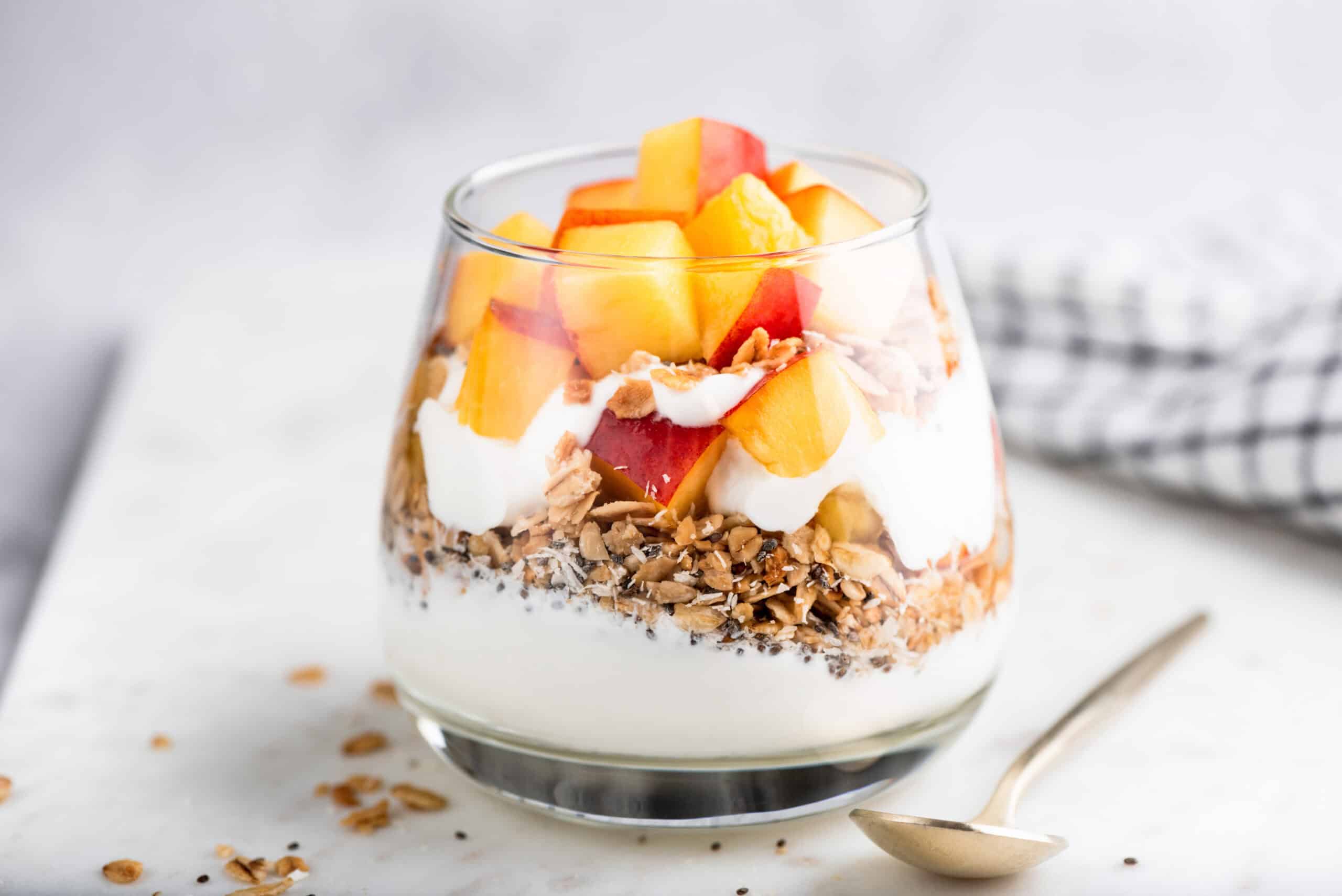 Glass of yogurt layered with granola and diced peach inclusions on a marble surface, showing how the right fruit piece shape delivers texture and appeal