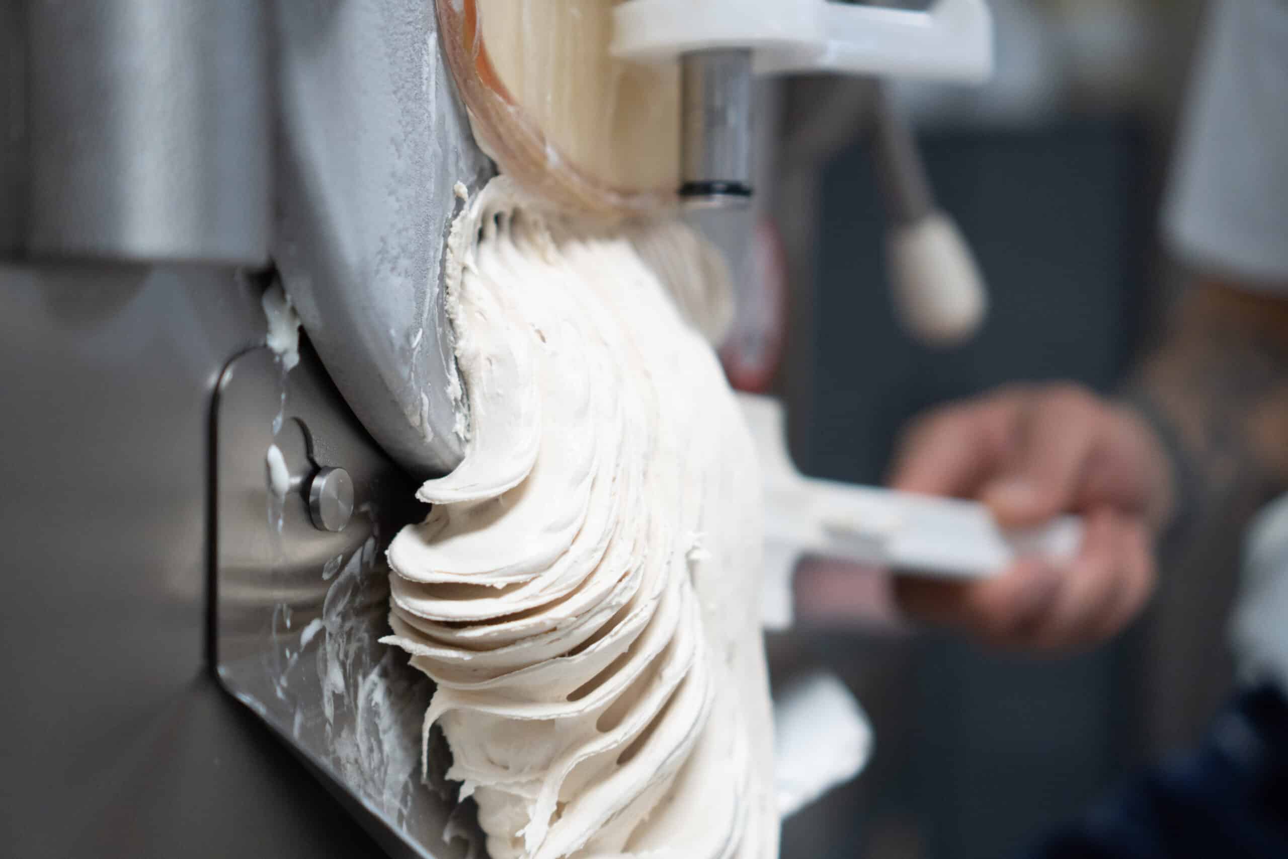 Soft ice cream base leaving a stainless steel batch freezer while a worker holds a spatula