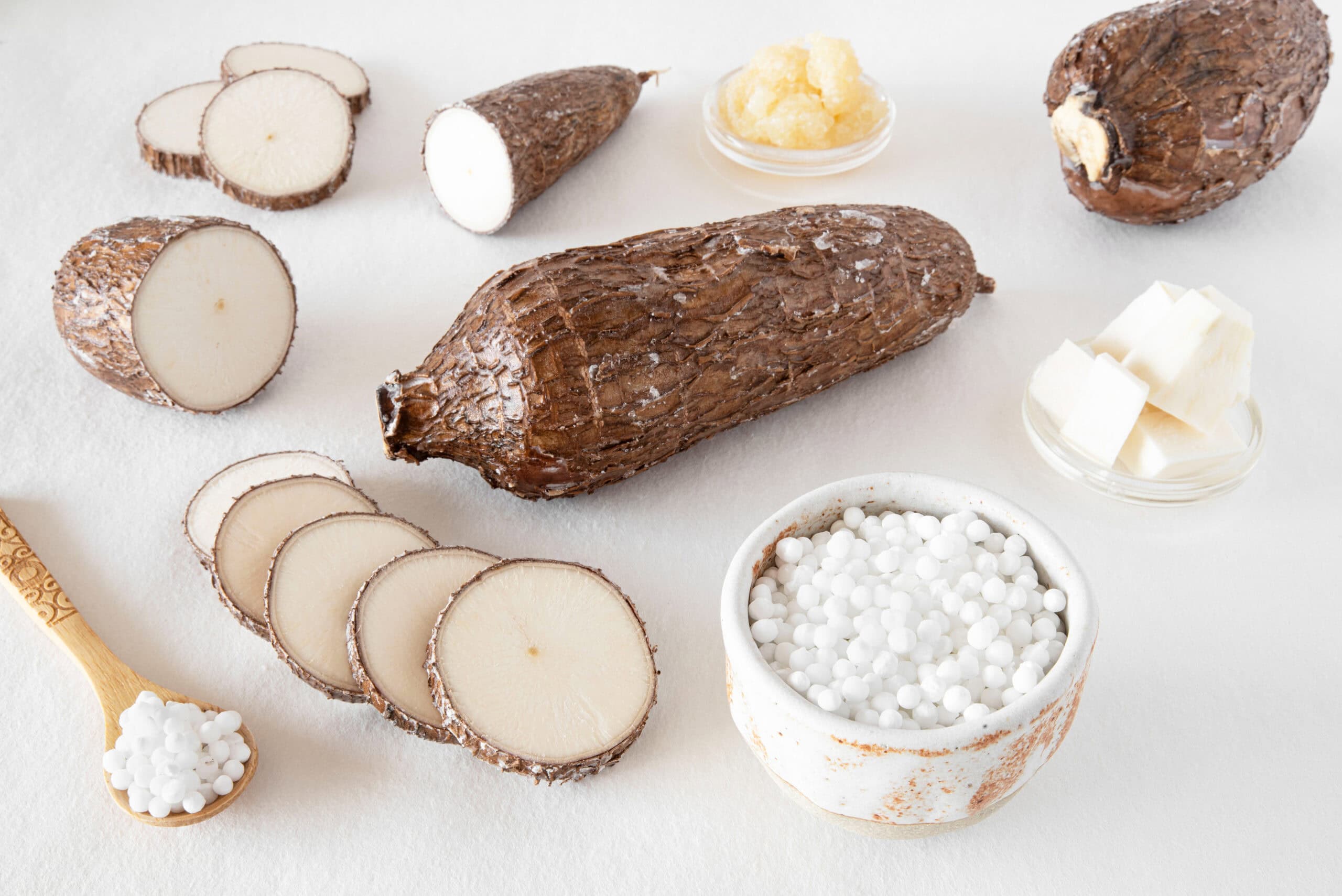 Clean label ingredients for texture: whole and sliced cassava roots beside bowls of tapioca pearls and prepared cassava on a white surface