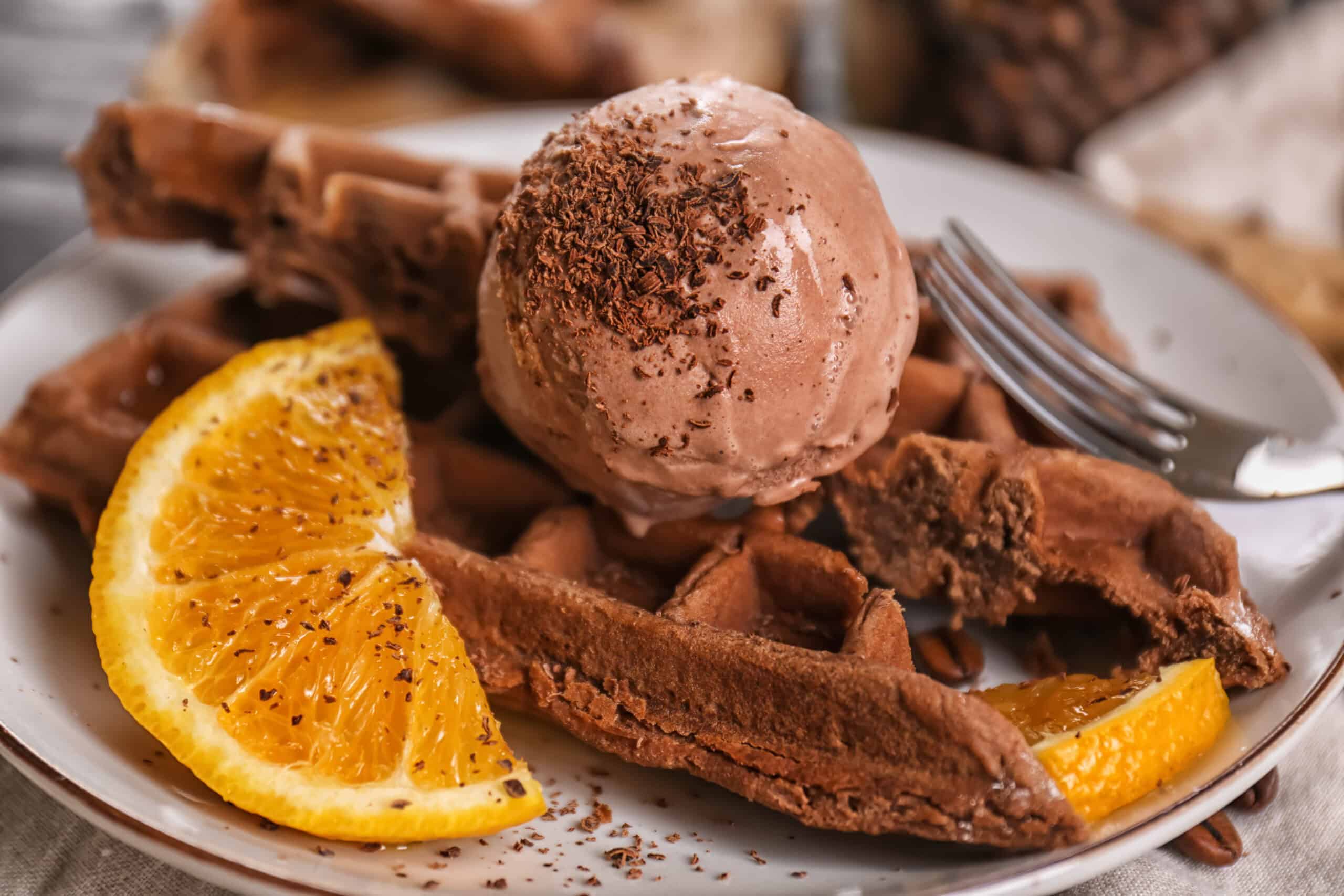Chocolate ice cream scoop topped with chocolate shavings on a chocolate waffle, beside orange slices on a plate