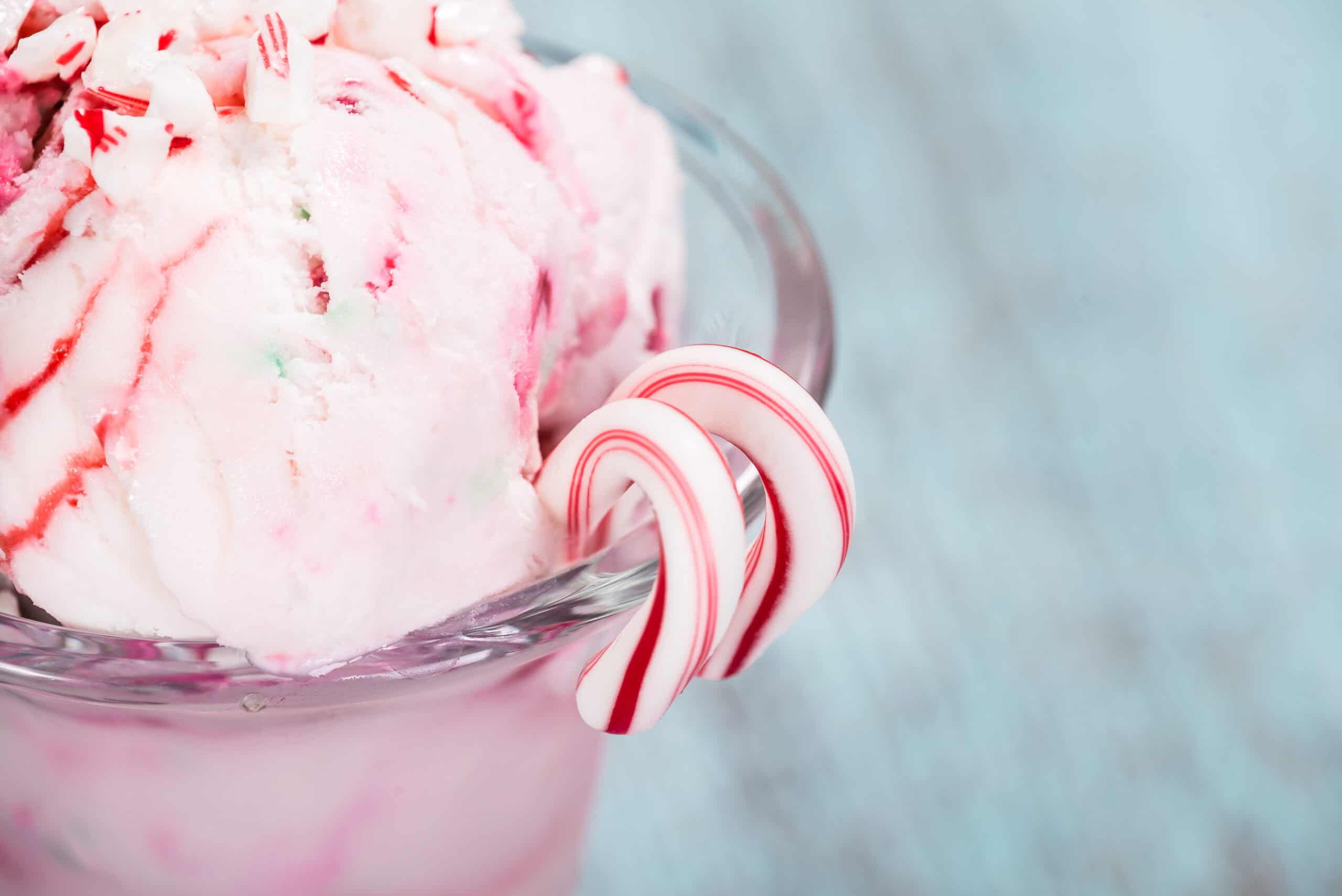 Peppermint ice cream scoop with crushed candy cane topping in a glass dish, with two mini candy canes on the rim