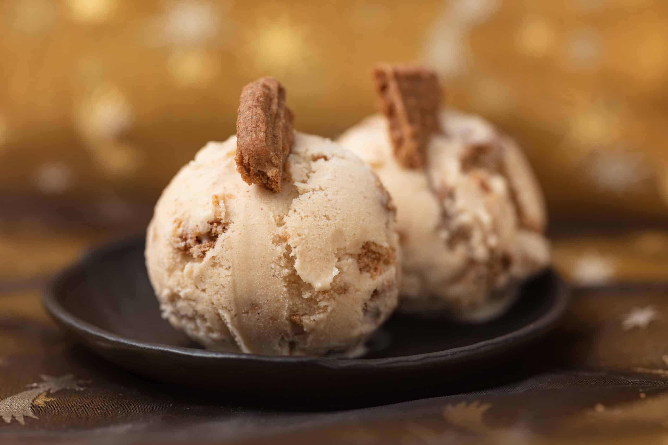 Two ice cream scoops topped with speculoos cookie pieces on a dark plate against a gold background