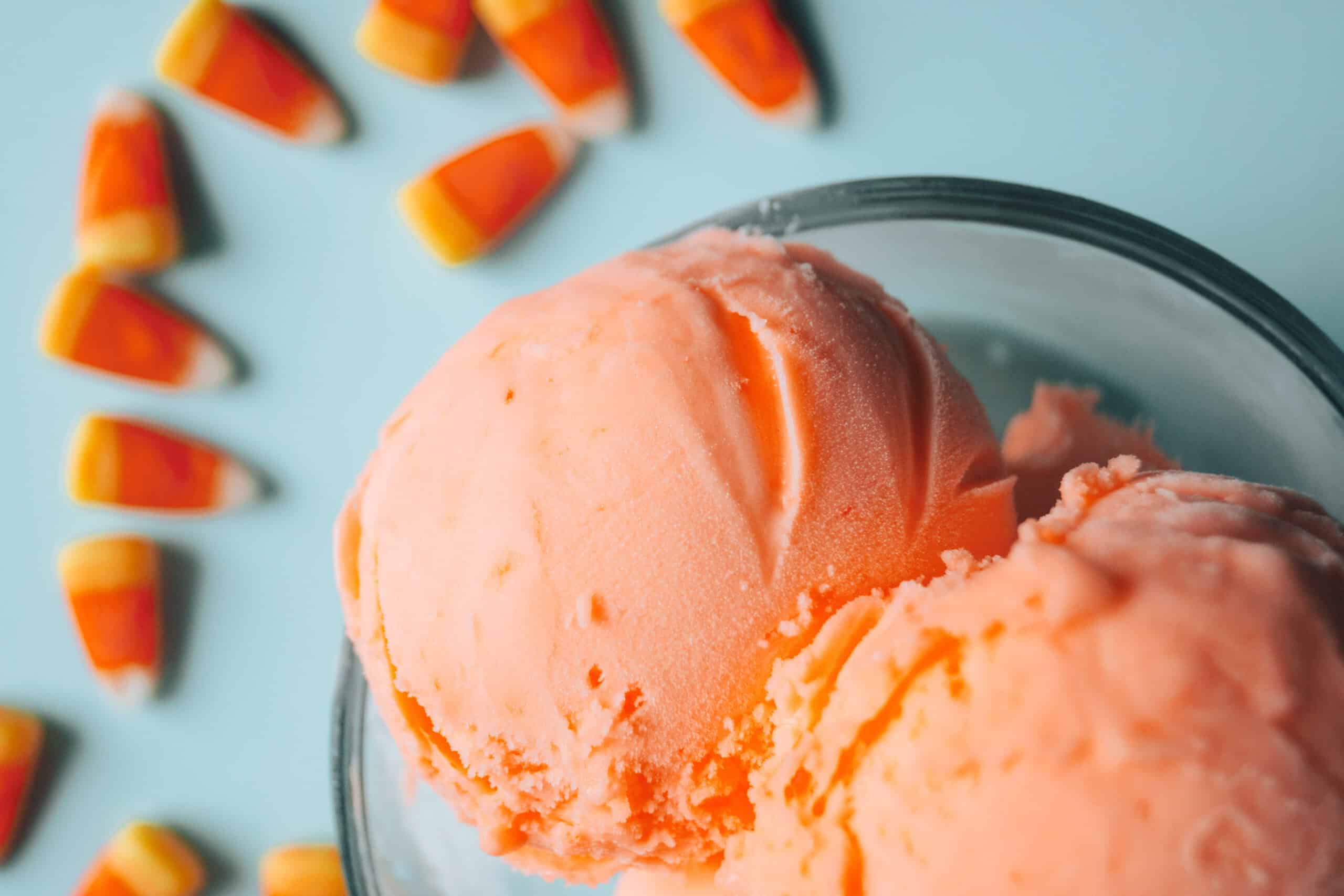 Close-up of orange ice cream scoops in a glass bowl with candy corn scattered on a light blue surface