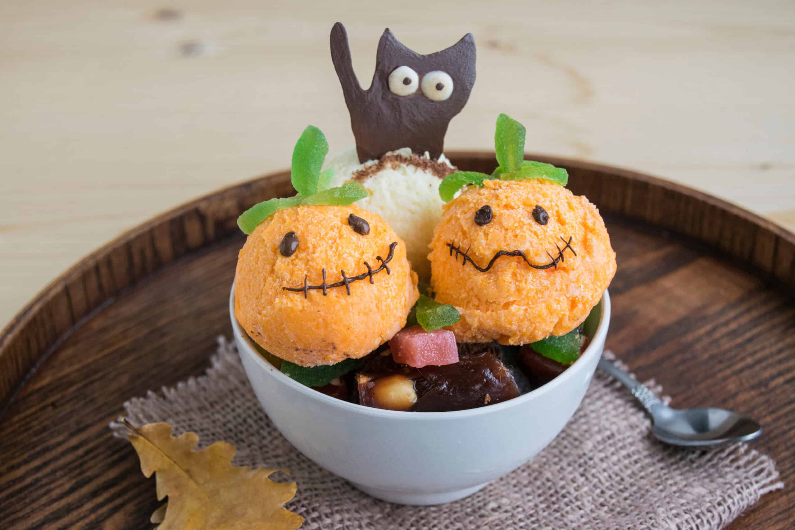 Two orange ice cream scoops with jack-o’-lantern faces beside a vanilla scoop and a chocolate cat cutout in a white bowl